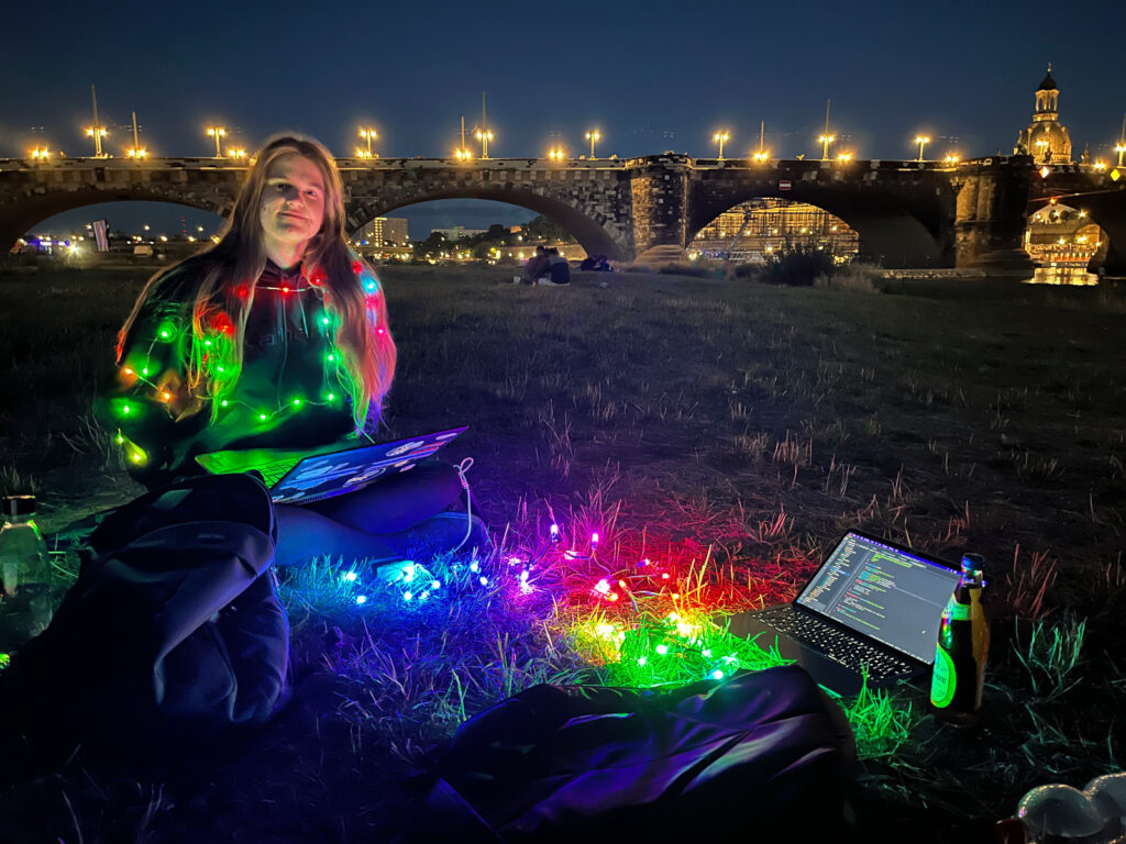 Frau sitzt auf Wiese mit schwarzem Pulli, welcher eine bunt leuchtende Lichterkette trägt. Auf ihrem Schoß ein Laptop. Auf der Wiese eine weitere Lichterkette und ein weiterer Laptop.